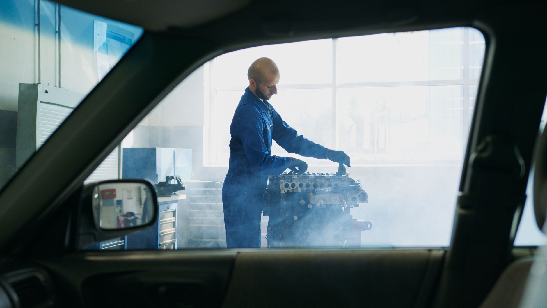 Engine on a stand giving off steam in a repair shop, seen through a car window